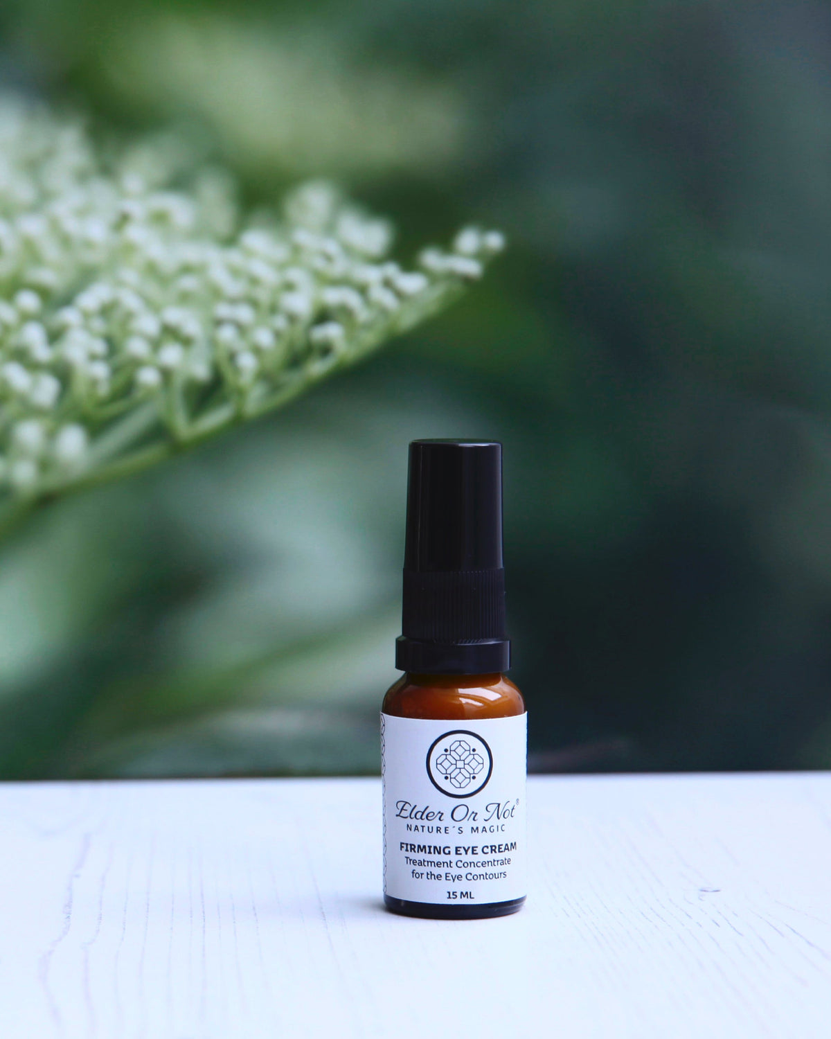 A small amber glass bottle of Firming Eye treatment concentrate with a black serum pump, standing on a white wooden surface with a soft-focus background of green leaves and elderflower buds.