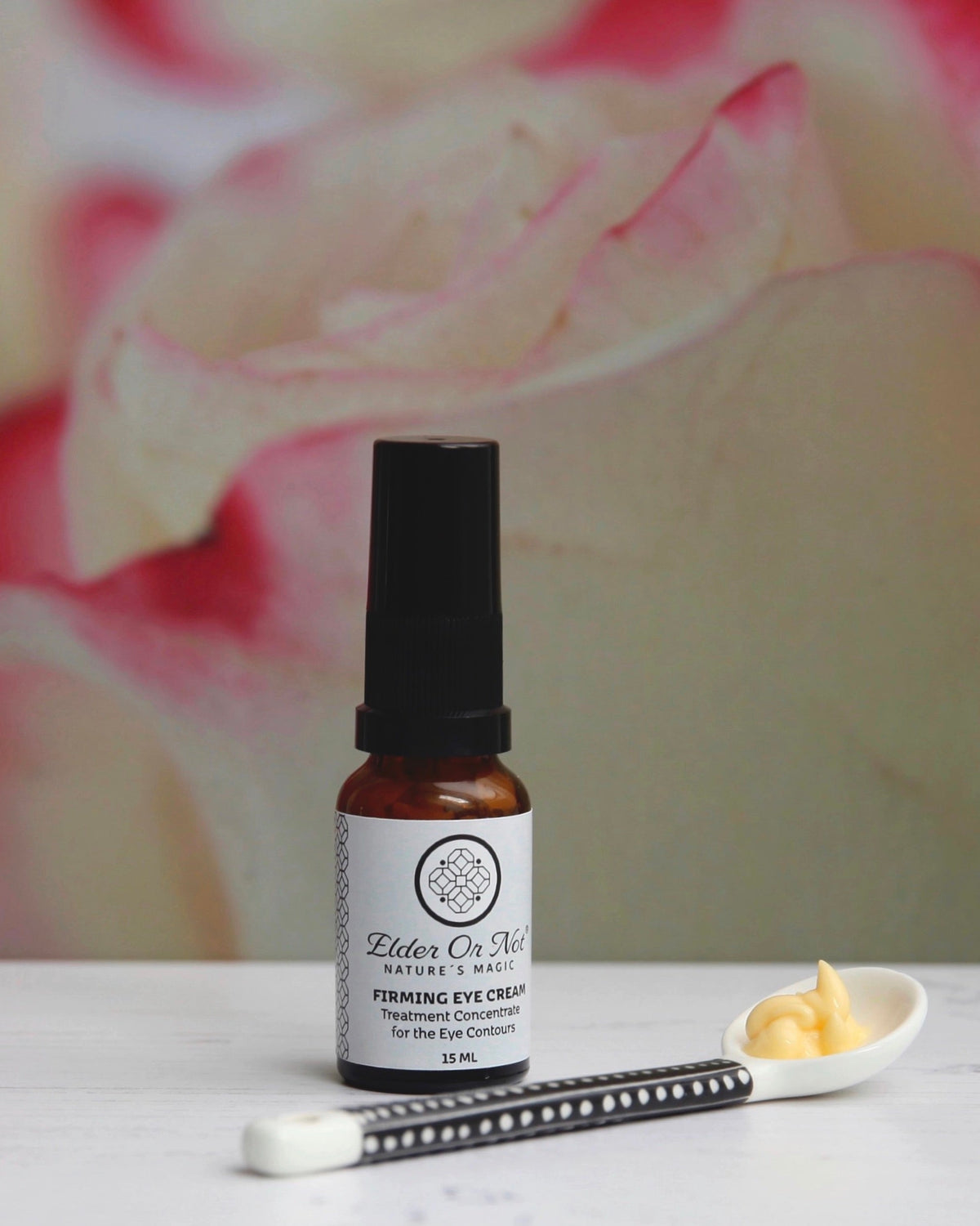A small amber glass bottle of Elder Or Not Skincare Firming Eye Cream with a black cap stands on a white surface. A small white spoon with yellow cream and a black-and-white applicator are placed in front, with a softly blurred pink rose in the background.