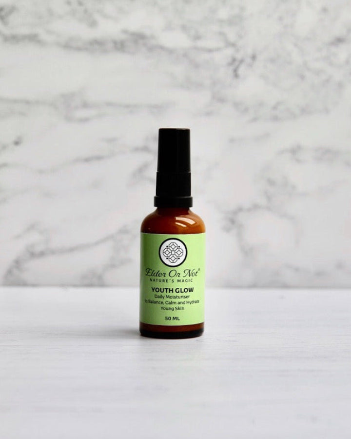A 50ml amber glass serum bottle with a black pump and an apple green label reading Elder or Not Skincare Youth Glow standing on a white marble surface against a white marble wall.