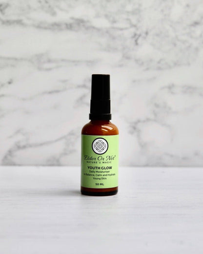 A 50ml amber glass serum bottle with a black pump and an apple green label reading Elder or Not Skincare Youth Glow standing on a white marble surface against a white marble wall.