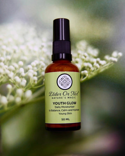 50 ml serum bottle of Youth Glow moisturiser in an amber serum bottle with black lid and an apple green label, with elderflower buds