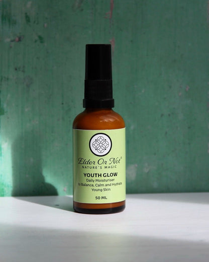 A 50 ml amber glass spray bottle of Elder Or Not Skincare Nature's Magic Youth Glow moirturiser, standing on a white surface against a textured green background.