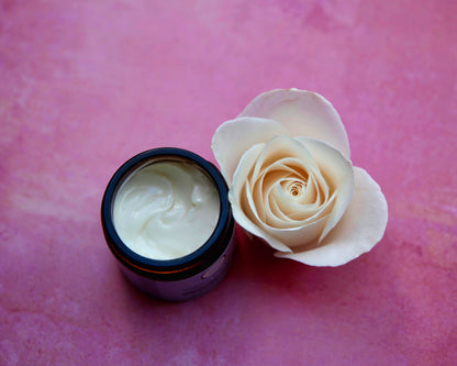 An open jar of white cream next to a cream-colored rose on a textured pink background.