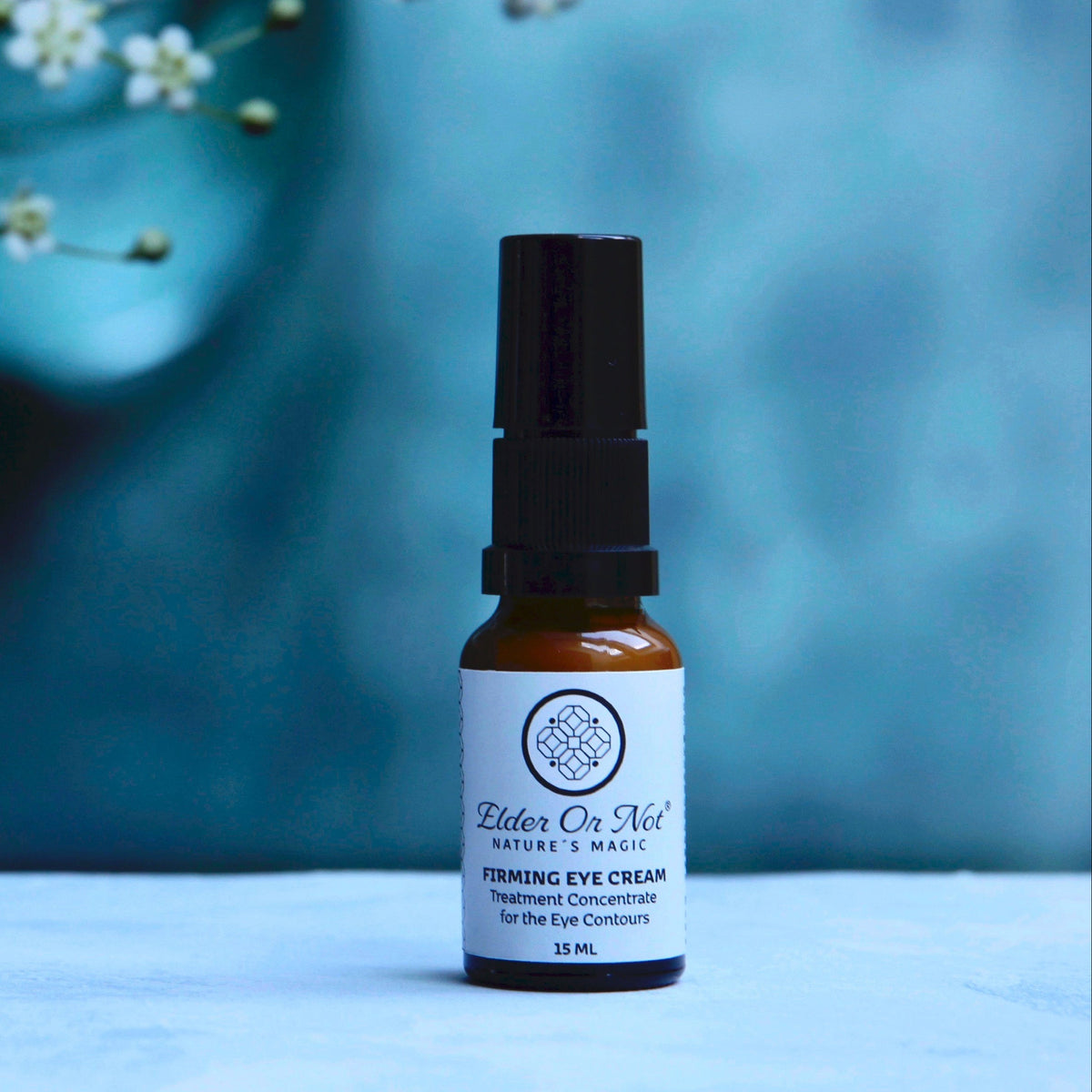A small amber glass bottle of Elder Or Nort Nature's Magic Firming Eye Cream with a black serum cap, set on a light marble surface against a textured blue background with white flowers.
