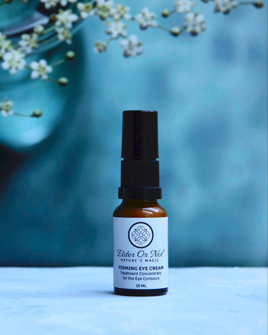 A small amber glass bottle of Elder Or Nort Nature's Magic Firming Eye Cream with a black serum cap, set on a light marble surface against a textured blue background with white flowers.