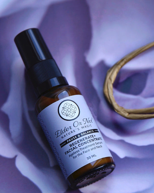 A small amber glass serum bottle with a black nozzle and a white label reading 'Elder Or Not Skincare' lies on a soft lavender-purple rose surface next to a gold-toned paper bag handle.