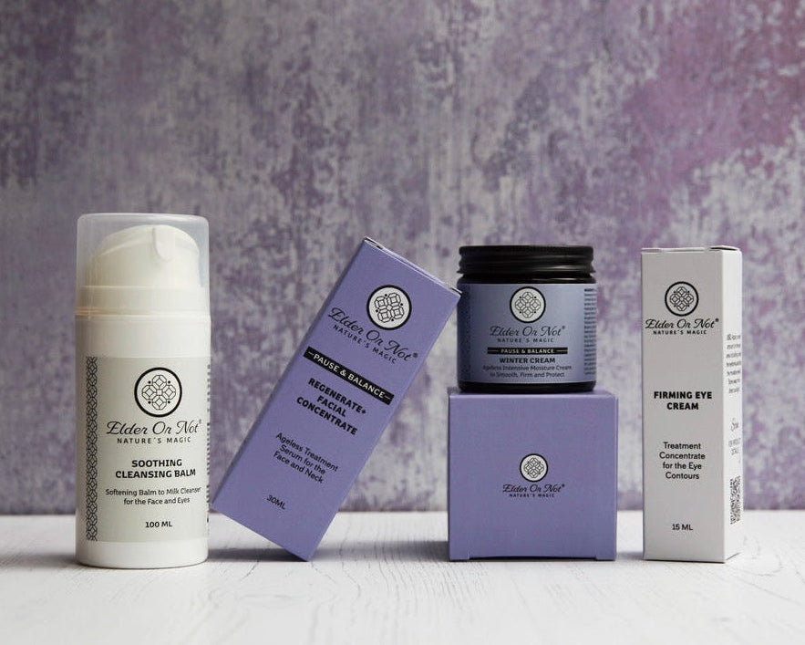 Four skincare products in the Winter Skincare collection from Elder Or Not Skincare arranged on a white surface against a textured frosty purple background: a white cleansing balm bottle, a purple box, a purple jar with a black lid on a purple box, and a white eye cream box.