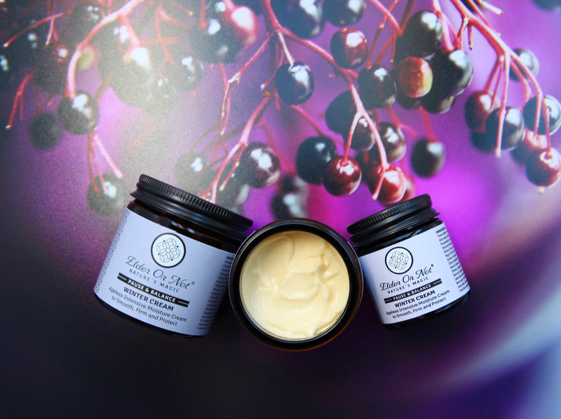 Two jars of winter cream with a container of cream in the center, set against a background of dark berries.