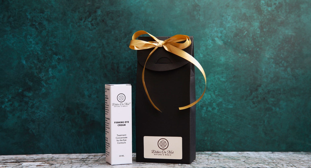 A white cosmetic eye cream bottle and a tall black gift box with a gold ribbon bow, both branded Elder Or Not Skincare set against a textured dark teal background.