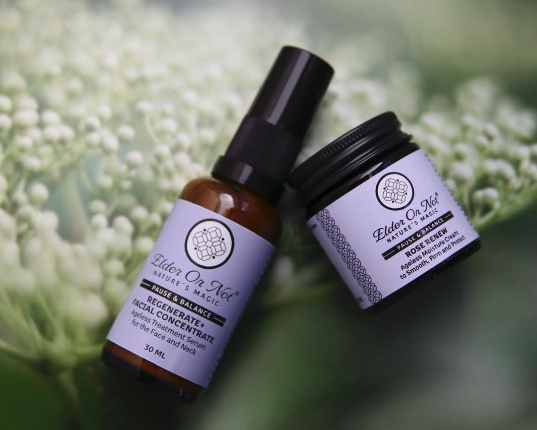 Two skincare products from Elder Or Not Skincare. One serum and a moisturiser both with lavender labels resting on a bed of elderflower buds.