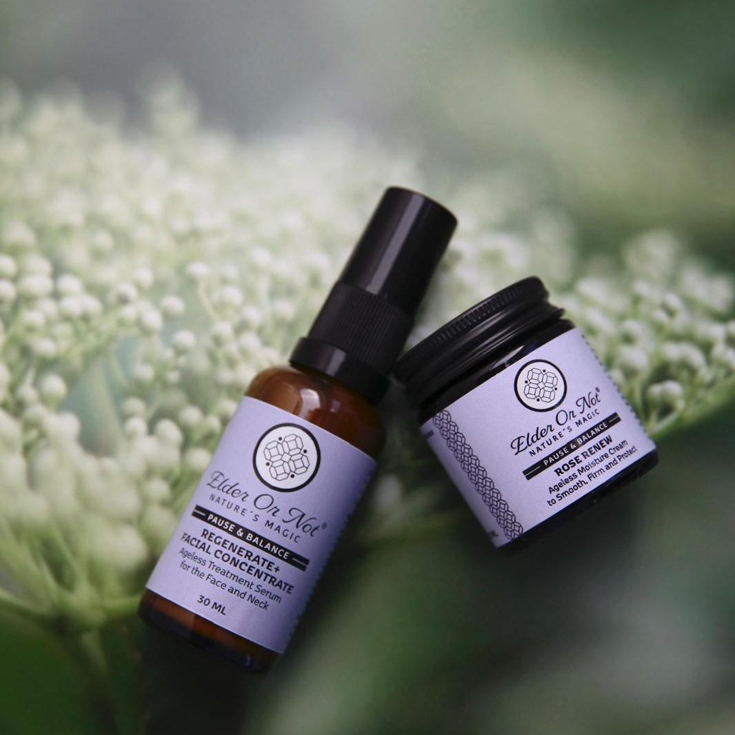 Two skincare products  30ml amber serum bottle and a small black jar of cream, both with lavender labels, resting on a bed of small white flowers with a soft green elderflower bud background.