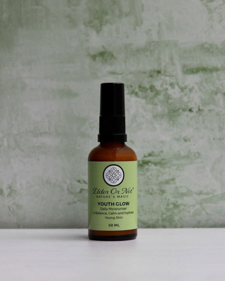 A small amber bottle with a black spray top and a light green label reading 'Balm of Nill Youth Colognatus' stands on a white surface against a textured pale green wall.