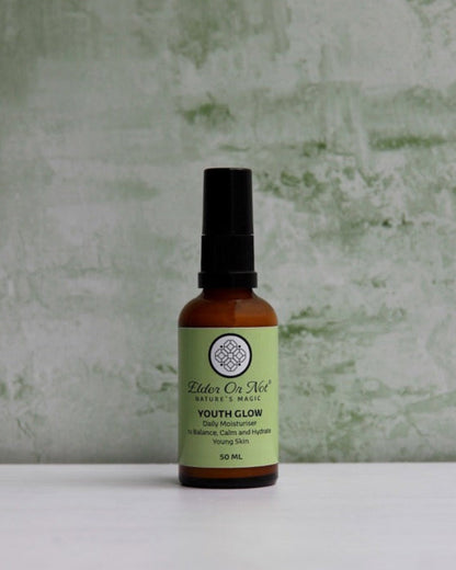 A small amber bottle with a black spray top and a light green label reading 'Balm of Nill Youth Colognatus' stands on a white surface against a textured pale green wall.