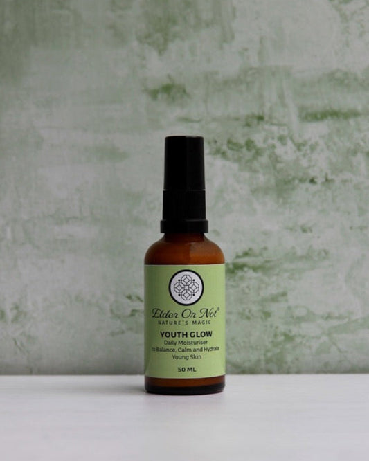 A small amber bottle with a black spray top and a light green label reading 'Balm of Nill Youth Colognatus' stands on a white surface against a textured pale green wall.